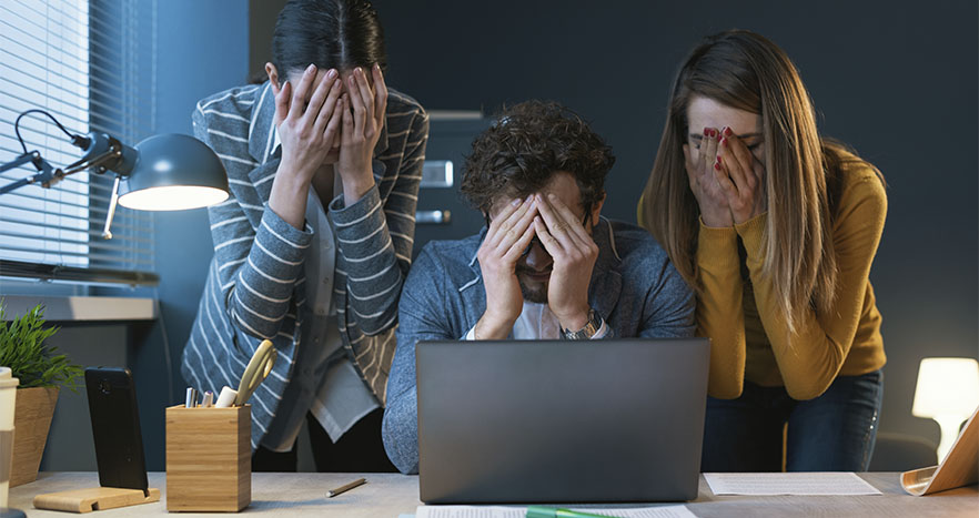 Três colegas de trabalho cobrindo seus rostos. À frente, um computador está aberto, indicando que houve algum com problema com a cibersegurança dos dados.