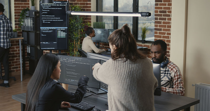 Programadora apontando para uma tela de computador, explicando a funcionalidade dos códigos aos seus colegas.